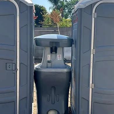 A portable handwashing station with a foot pump, standing between two grey portable toilets on a jobsite