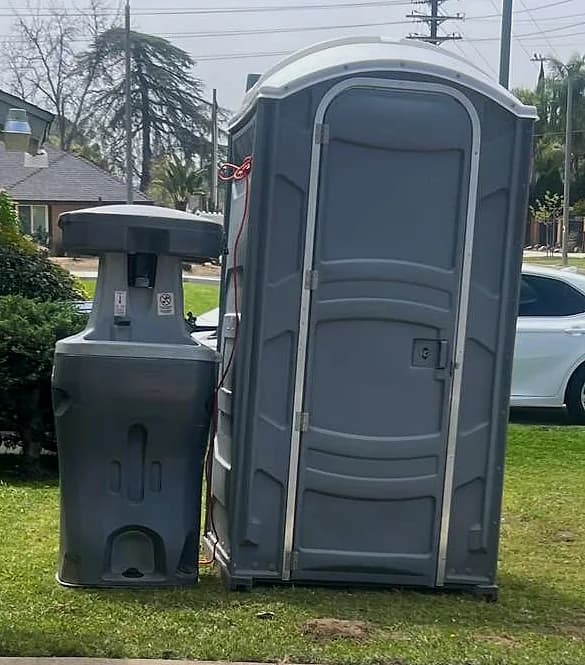 A portable handwashing station with a foot pump standing beside a grey portable toilet on cut grass