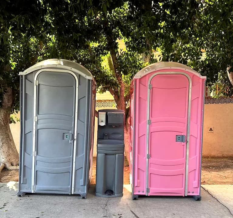 A pink portable toilet beside a handwashing station and a grey portable toilet, placed on a shaded patio