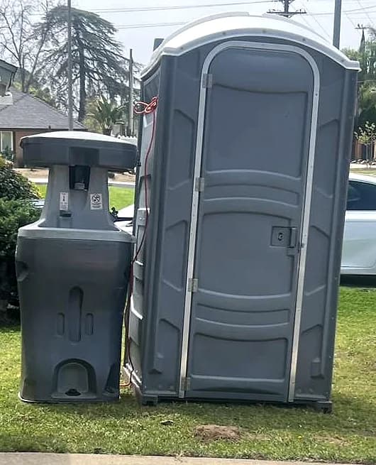 A complete grey portable toilet standing beside a portable handwashing station on cut grass