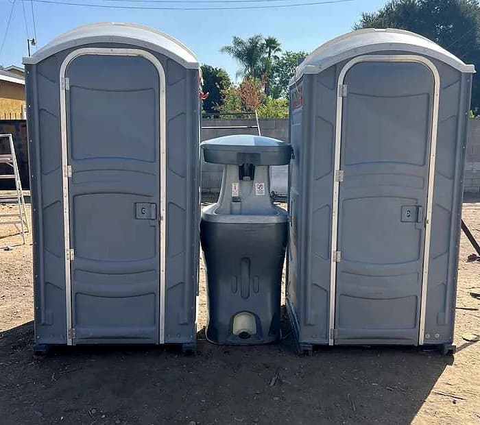 Two portable toilets flanking a portable handwashing station on a graded dirt jobsite