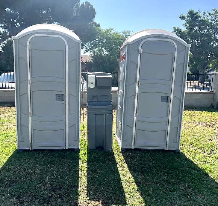 Two standard grey portable toilets standing either side of a portable handwashing station on cut grass